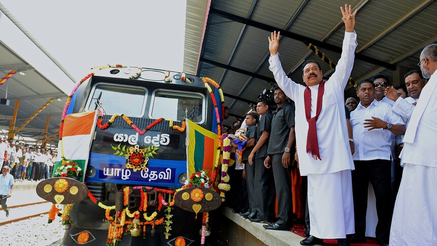 From the train tracks, you see a stationary elaborately-decorated train while Mahinda Rajapaksa stands on the platform beside it
