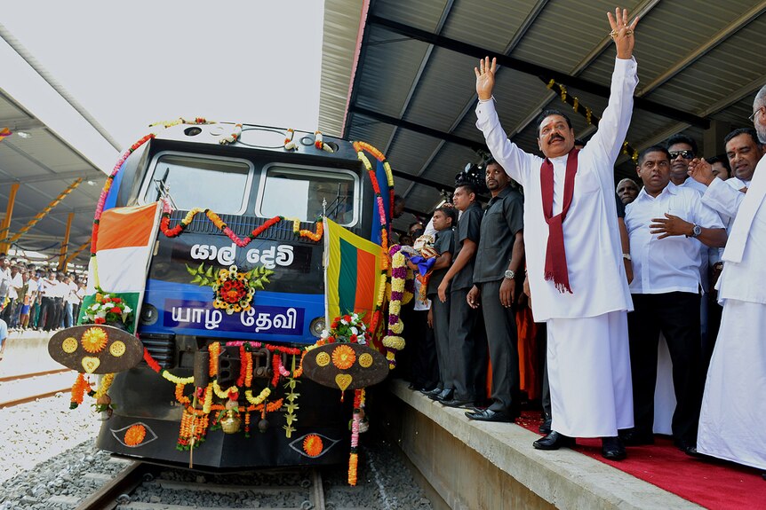 From the train tracks, you see a stationary elaborately-decorated train while Mahinda Rajapaksa stands on the platform beside it