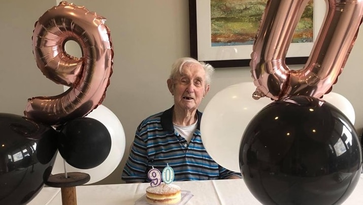 Alf Jordan sits at a table smiling, surrounded by balloons a small cake with candles in the shape of the number 90.