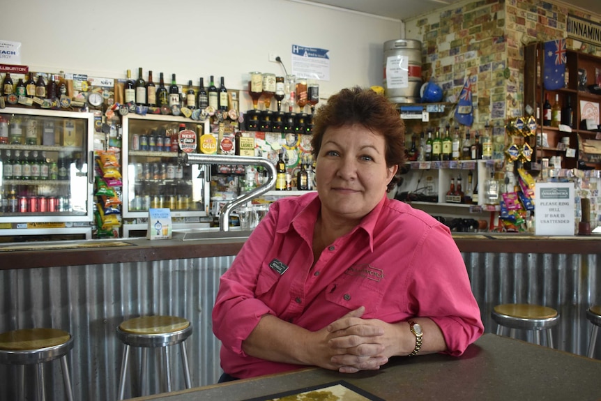 A lady in a pink shirt sits at a bar table.  She has blue eyes and is looking at the camera