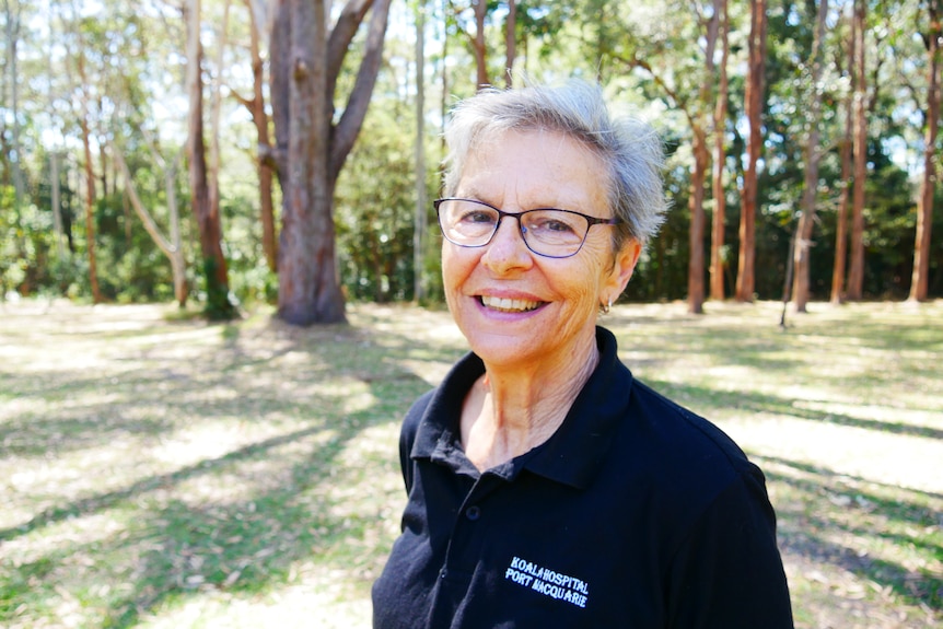 A woman with short grey hair and glasses smiles with trees behind her.