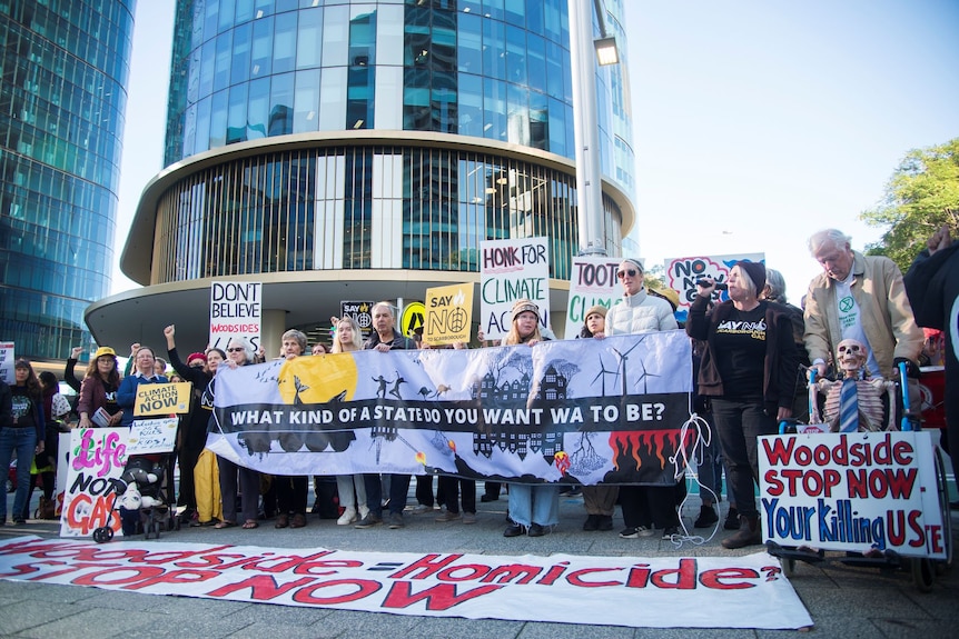 A group of protesters holding banners and placards stand outside a glass office building.