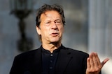 Imran Khan wearing a black shirt speaks as he raises his left hand up palm upwards