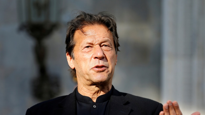 Imran Khan wearing a black shirt speaks as he raises his left hand up palm upwards