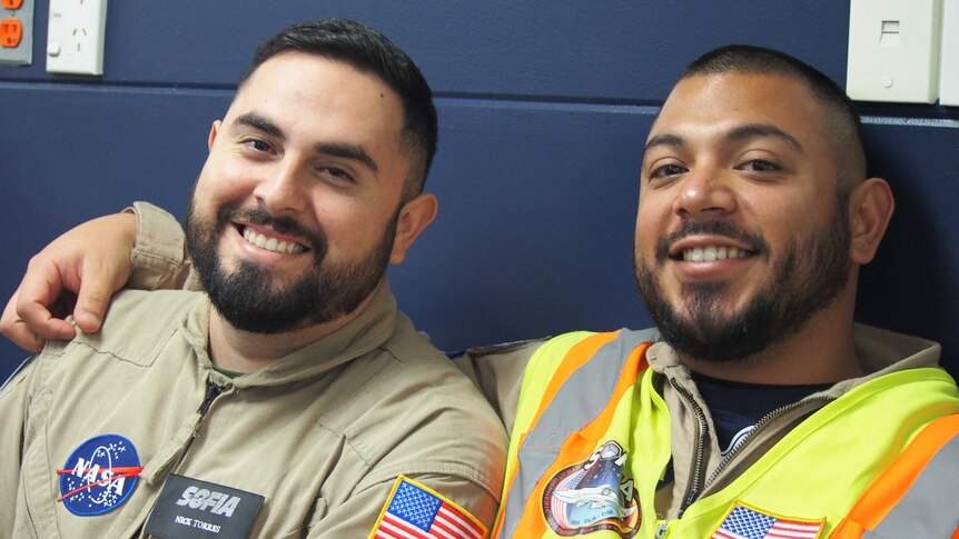 Two men smiling and wearing outfits badged with NASA symbols and the American flag.