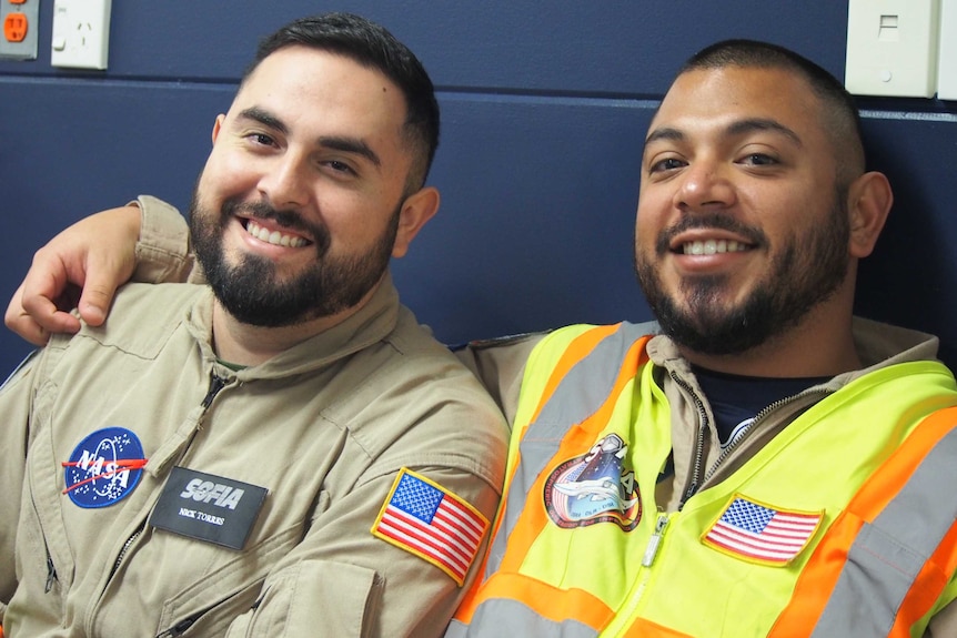 Two men smiling and wearing outfits badged with NASA symbols and the American flag.