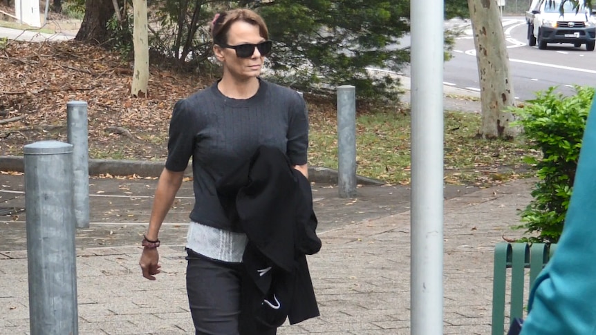 A woman wearing dark sunglasses and dark clothing walks along a footpath.