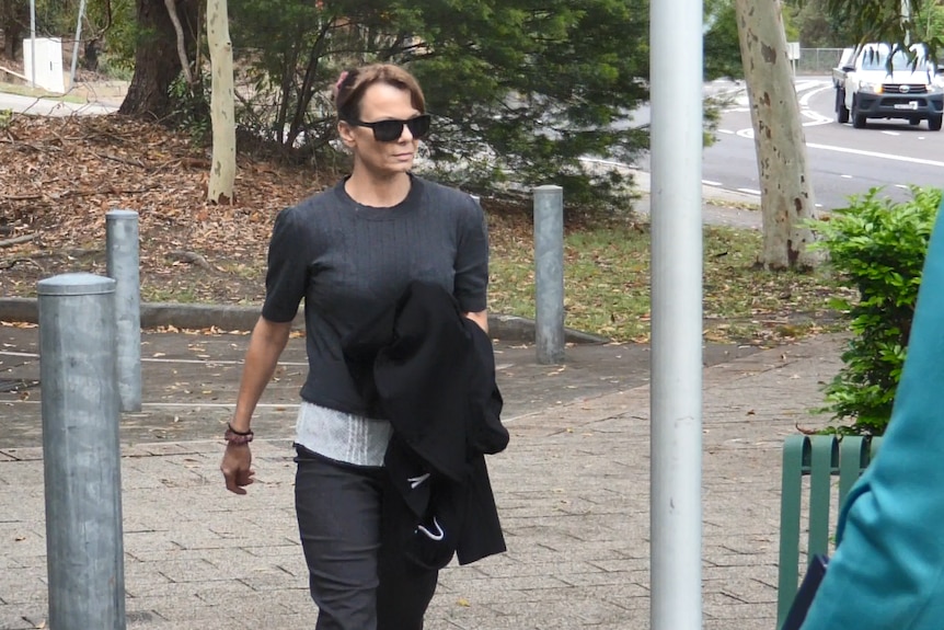 A woman wearing dark sunglasses and dark clothing walks along a footpath.