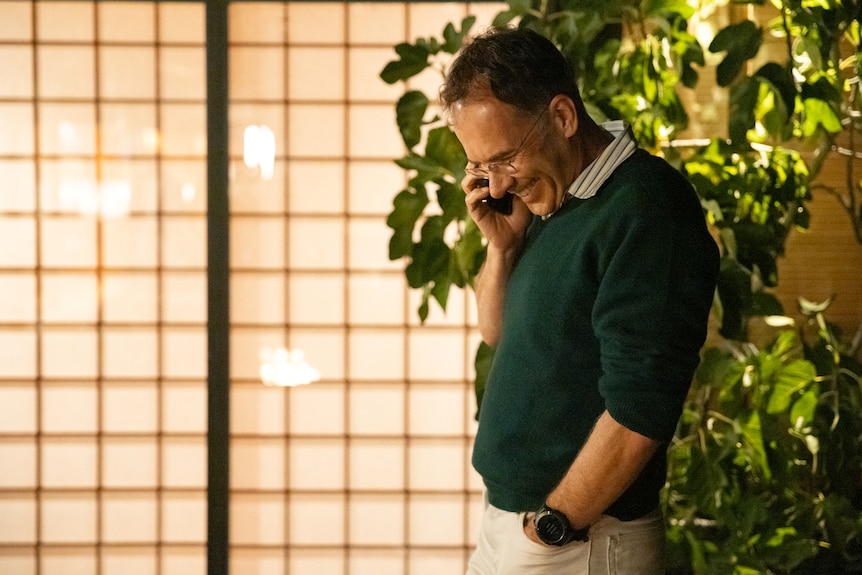 A man in a green jumper smiles as he looks down while talking on the phone in his home.