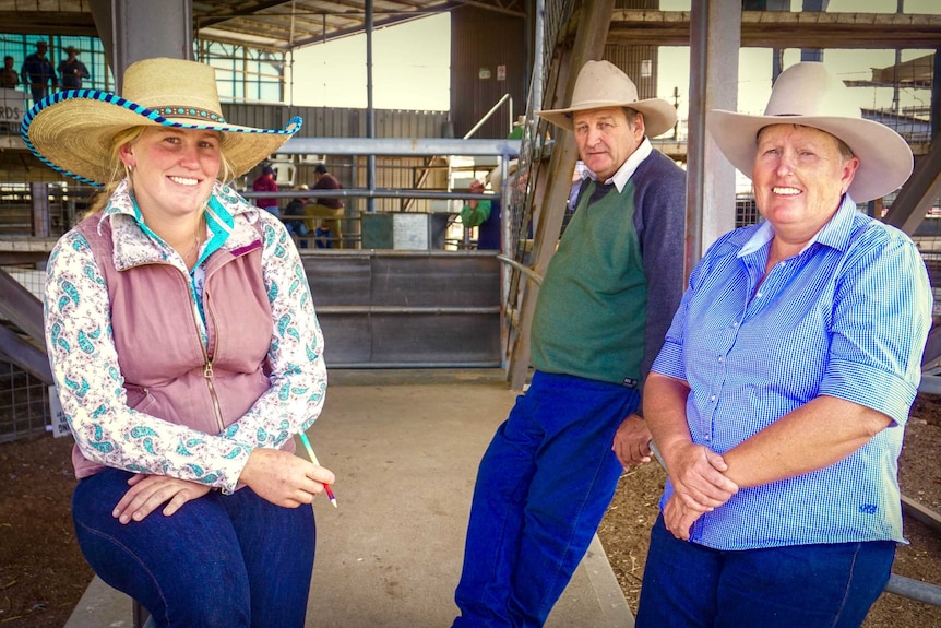 Three rural people in hats stand in a cattle yard smiling at the camera