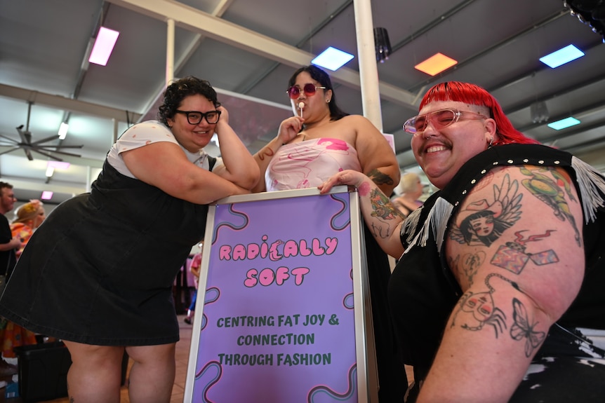 Three people pose next to a purple sign that reads ‘Radically Soft, centering fat joy and connection through fashion’. 