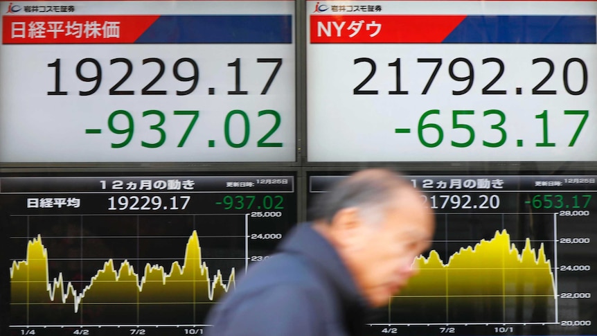 An old man walks past Japanese Nikkei indexes shown on screens who is captured mid-motion.