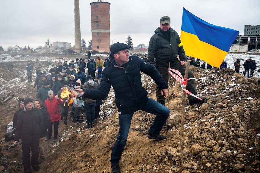 man in a black jacket and jeans stands on a hill with a ukrainian flag and people behind him prepares to throw molotov cocktail