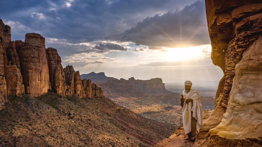 A priest in white robes stands outside the entrance to a cave in the desert.
