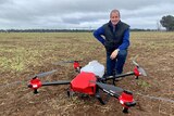 A man kneels behind a large drone with a spray container on the back in a paddock. 