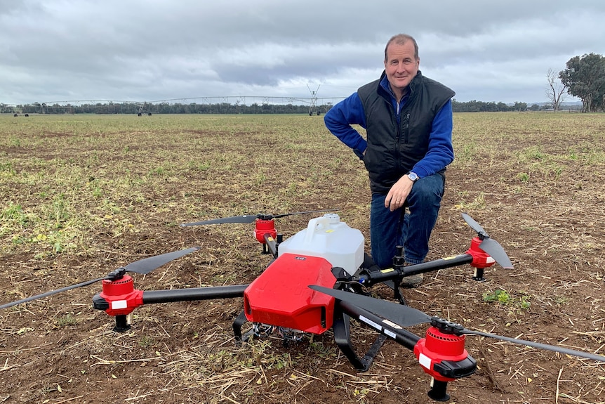 A man kneels behind a large drone with a spray container on the back in a paddock. 