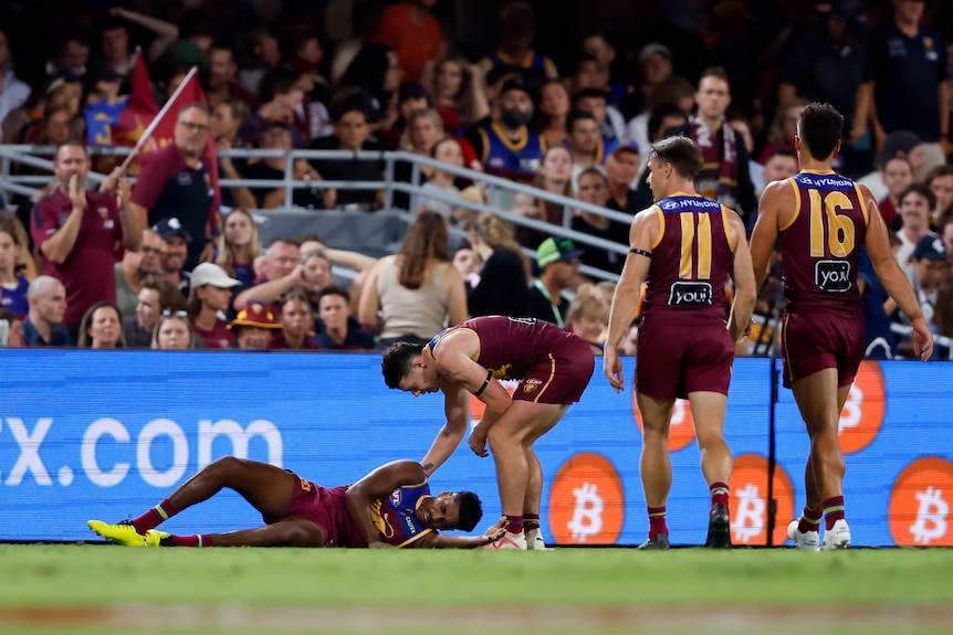 A Brisbane Lions player lies on the ground as his teammates gather round him.