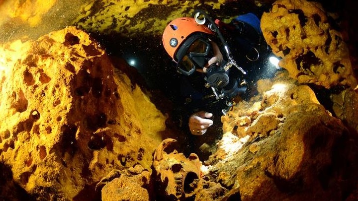 A scuba diver is surrounded by rock as they looks at an animal skull at Sac Actun underwater cave system.