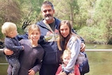 A bearded man posing with four children of varying ages including a toddler, in front of a river.