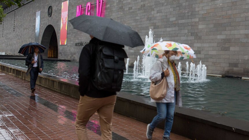 Τρεις άνθρωποι περνούν δίπλα από το NGV με ομπρέλες στη βροχή.
