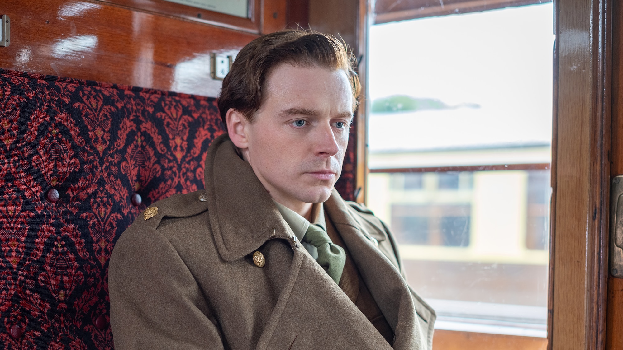 White man with short dark blonde hair wears taupe 1940s trenchcoat in a train carriage with maroon seats.