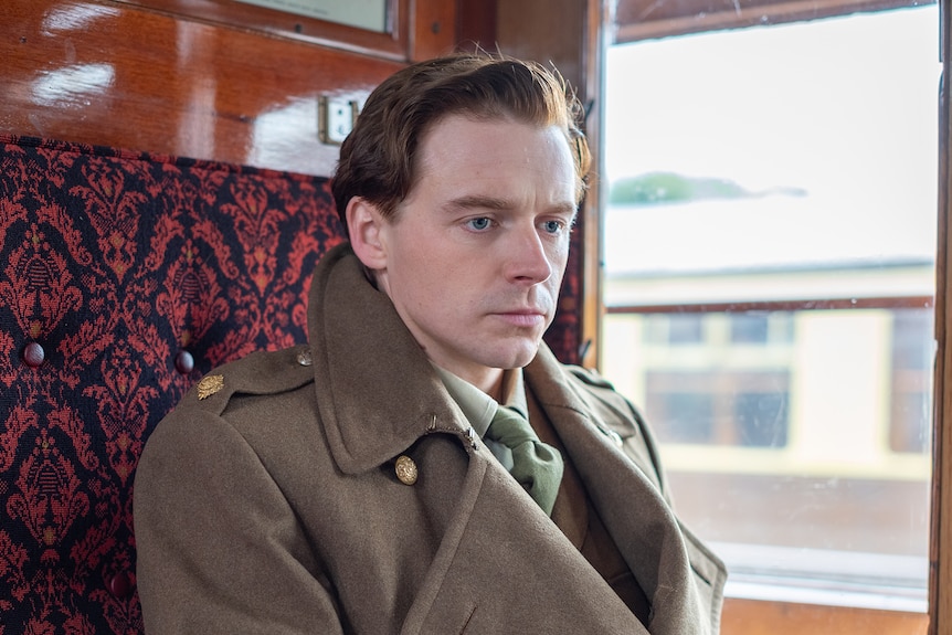 White man with short dark blonde hair wears taupe 1940s trenchcoat in a train carriage with maroon seats.