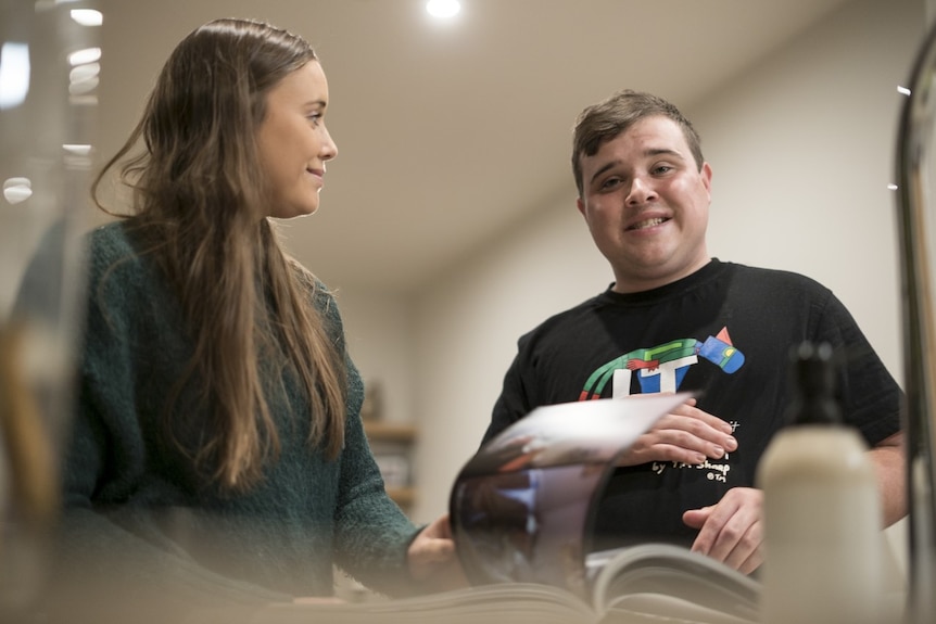 Tom Neale smiles at the camera with his sister standing next to him in their family home