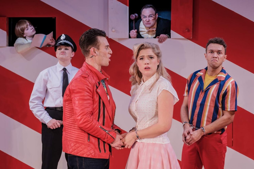 Production still showing six actors in 50s-style clothing on a colourful pink and red set.