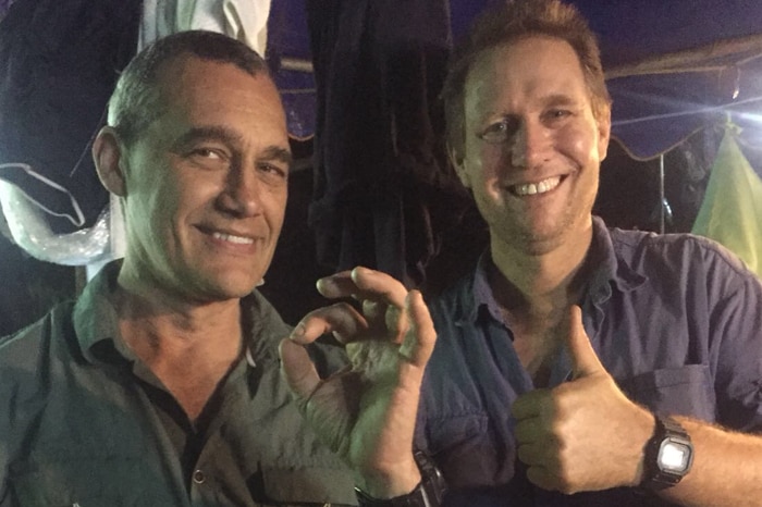 Two men in dark short sleeved shirts give a thumbs-up signal in a dimly lit room