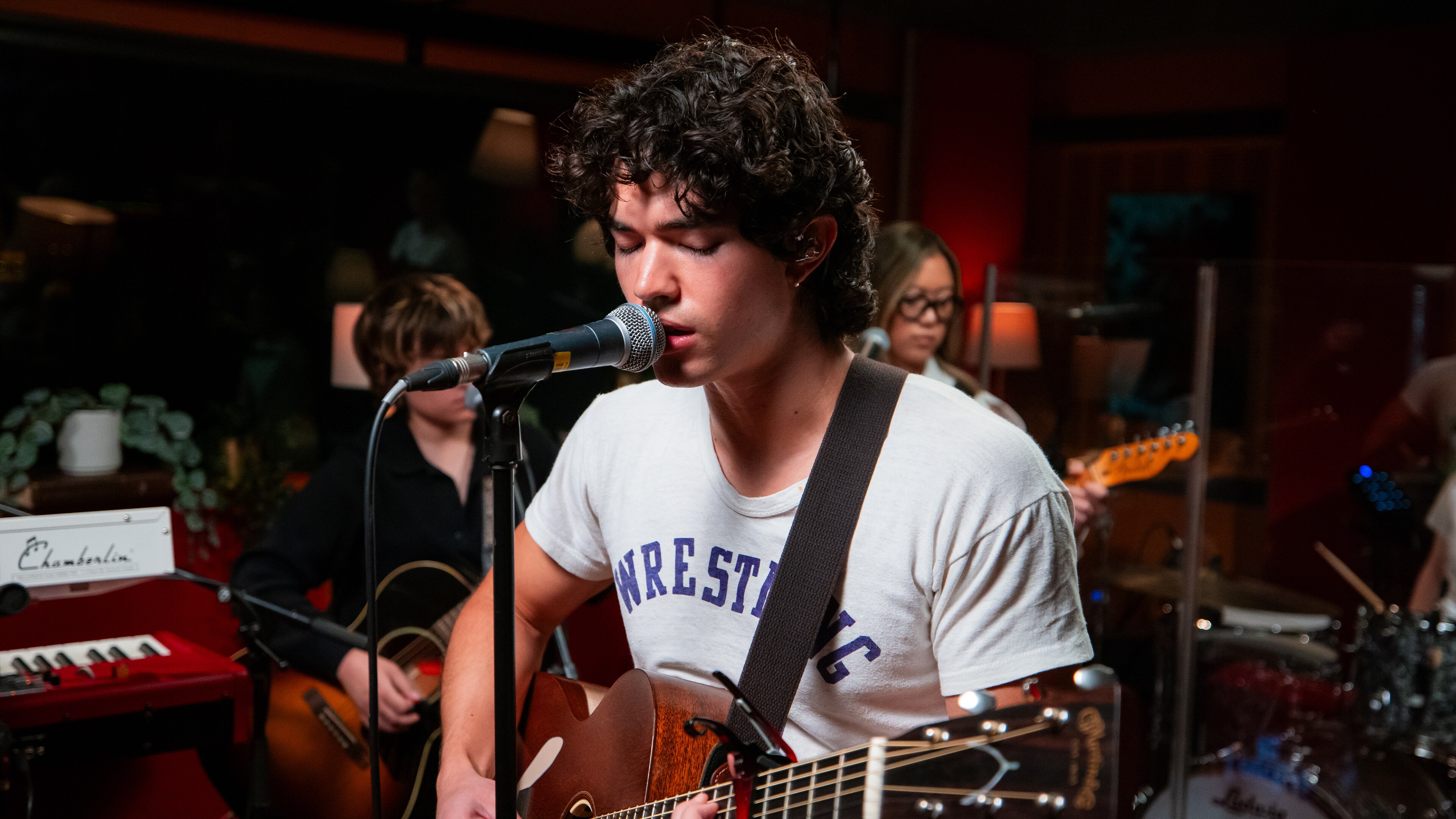 Conan Gray, wearing a white t-shirt, strums an acoustic guitar and sings into a microphone.
