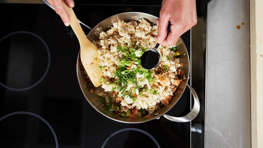 A pan full of rice with hands stirring with a wooden spoon and adding soy sauce.