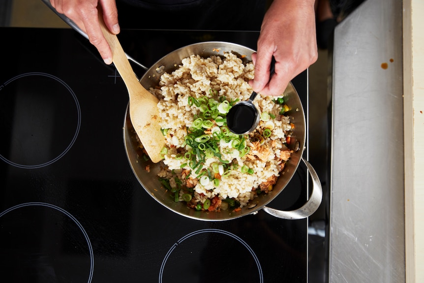 A pan full of rice with hands stirring with a wooden spoon and adding soy sauce.