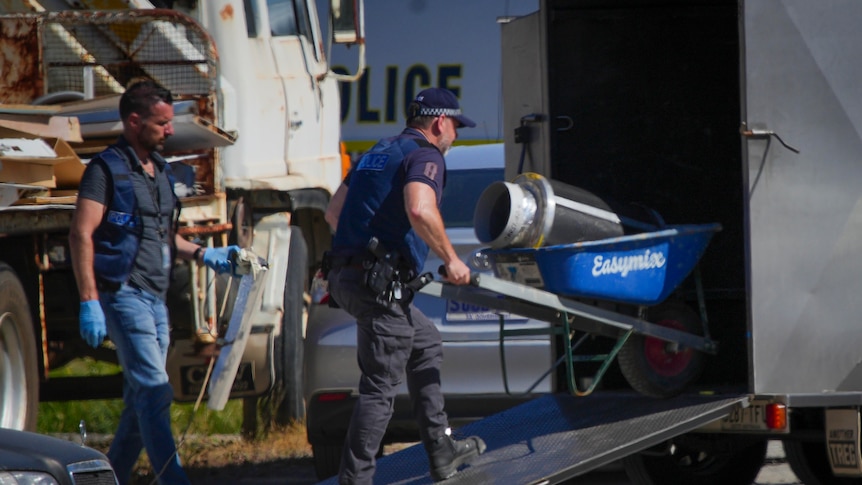 A police officer wheels a wheelbarrow with a large round metal object in it up a ramp into the back of vehicle