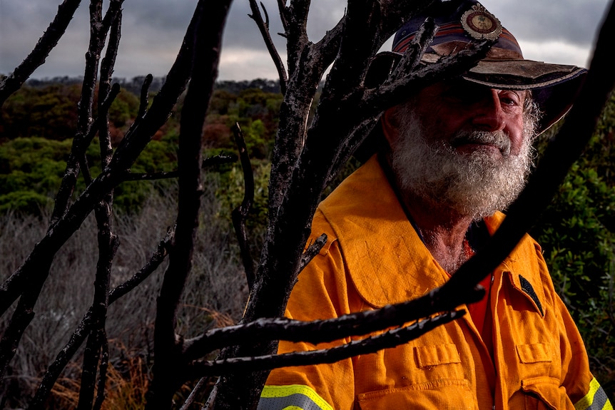 man in 70s with grey beard wearing fire uniform, standing next to tree with trees in the background