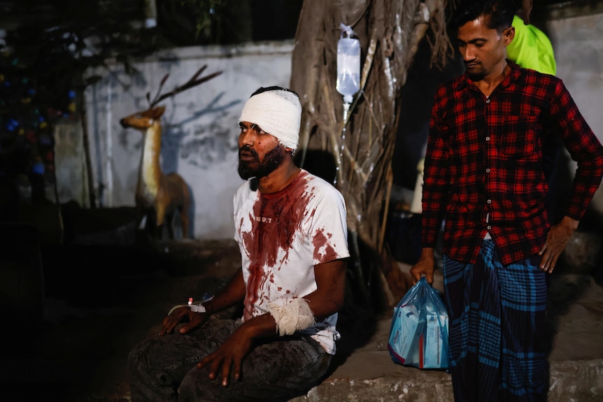 A man sits in a bloodied shirt with bandaging around him, another man behind him.