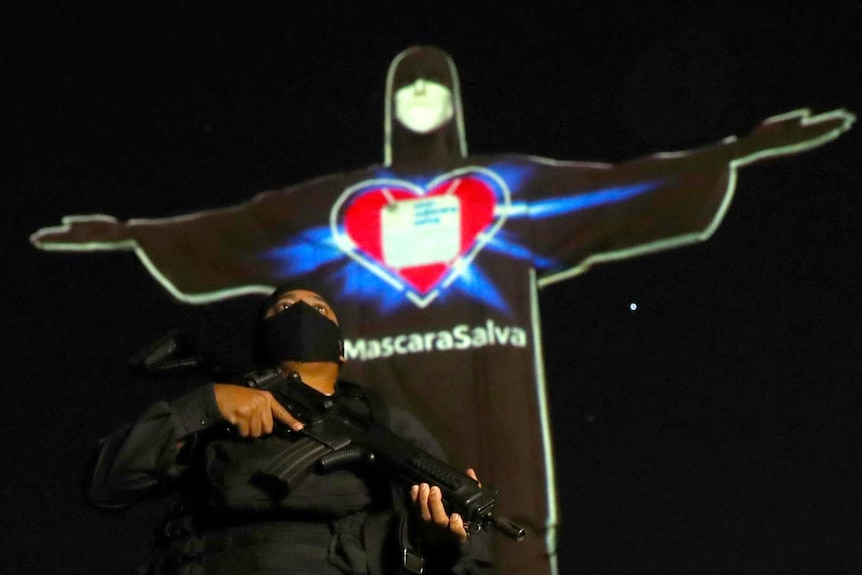 A uniformed police officer with a gun stands in front of the Christ the Redeemer statue, which has been lit up with a face mask