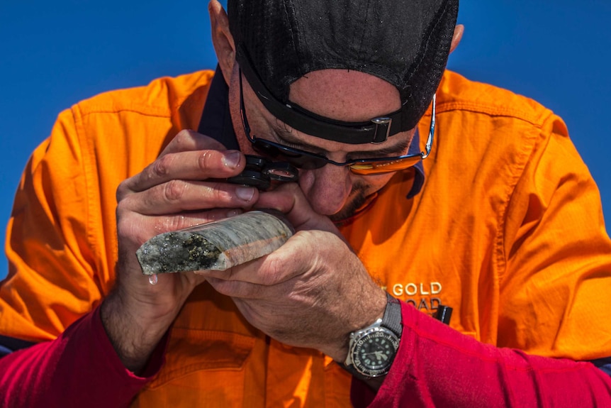 A man in a high-vis shirt using a magnifying device to look close up at samples of rock.