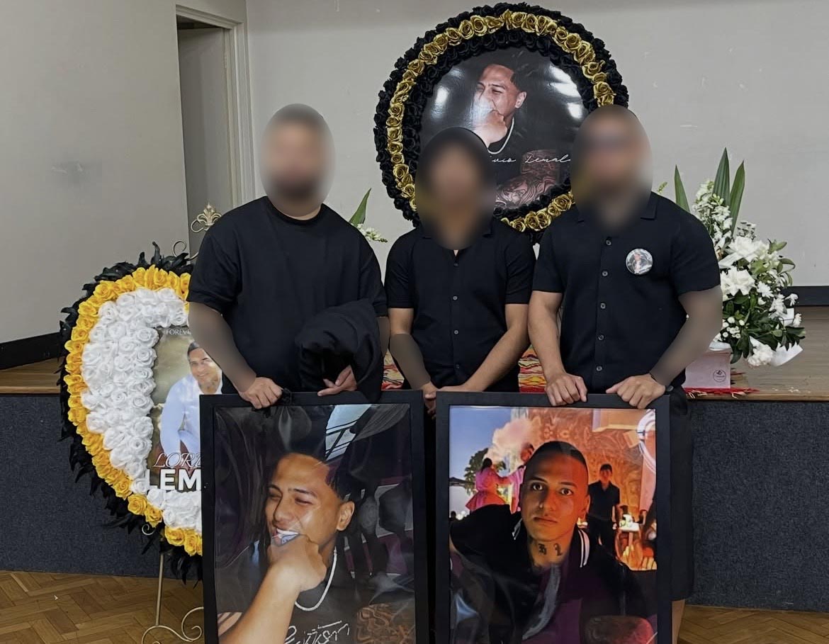 Three people with faces and tattooed arms blurred stand behind large image of man with shaved head, flowers behind them