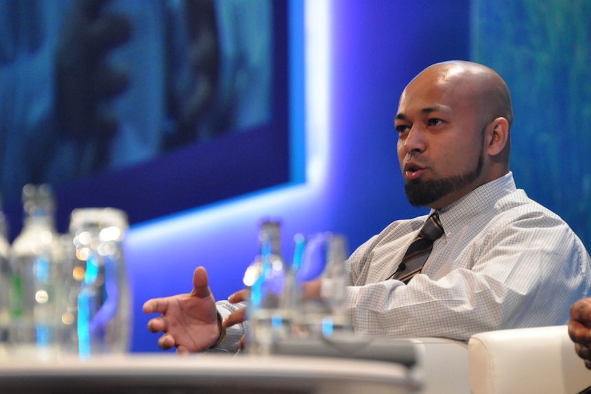 Man sits at a conference table wearing a white button up shirt and a tie. 