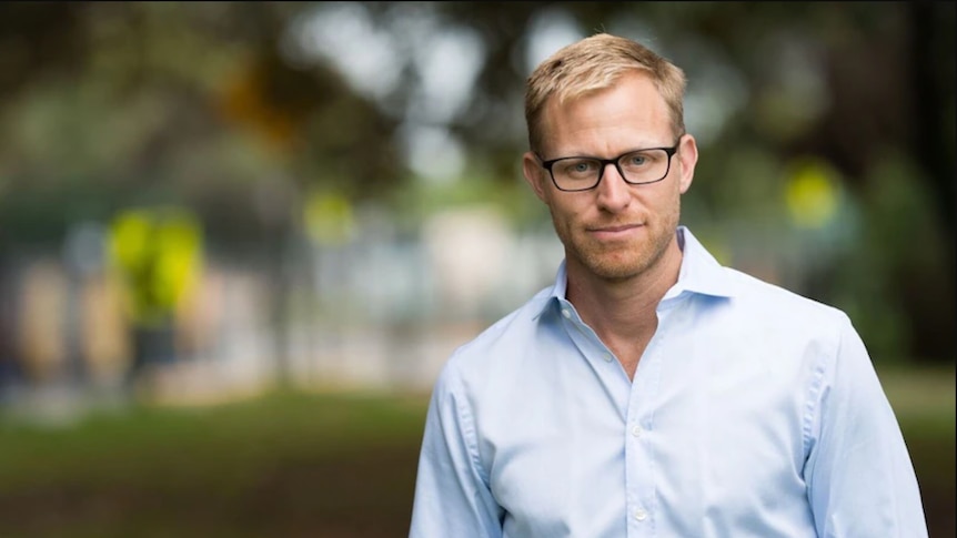 A fair-haired, bespectacled man in a light-coloured business shirt stands in a park.