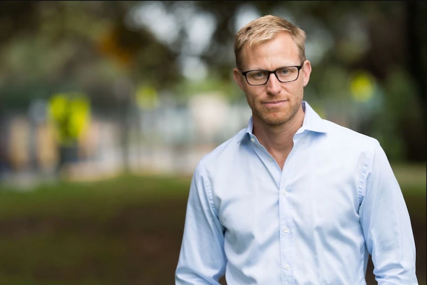 A fair-haired, bespectacled man in a light-coloured business shirt stands in a park.