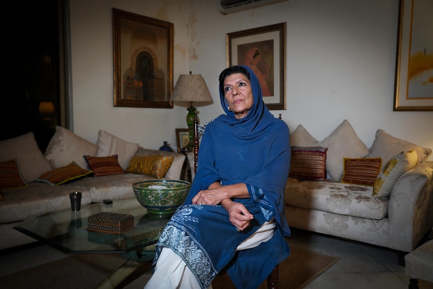 A woman wearing a blue head scarf sits on a glass table in a living room