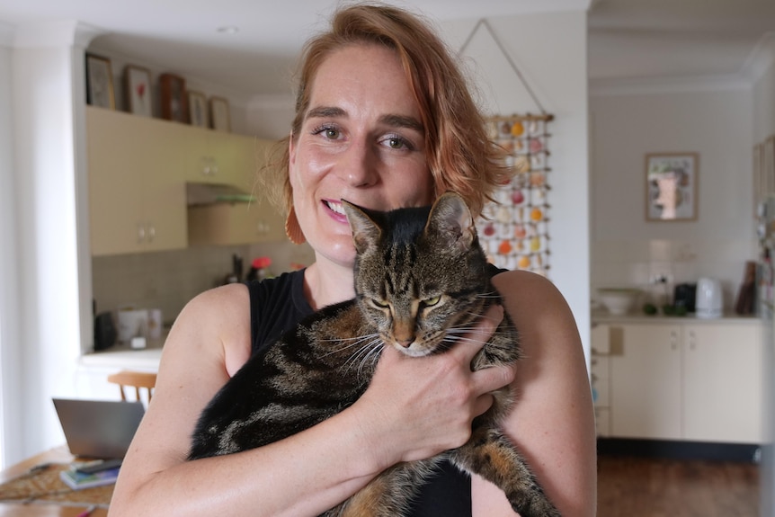 A woman holds her tabby cat in a cuddle to her chest, smiling in her home.