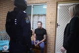 A man sitting outside a house with an armed policeman in front of him.