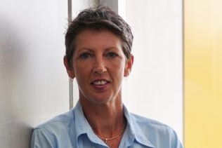 Dr Jo Coghlan with short brown hair standing against a wall wearing a blue shirt