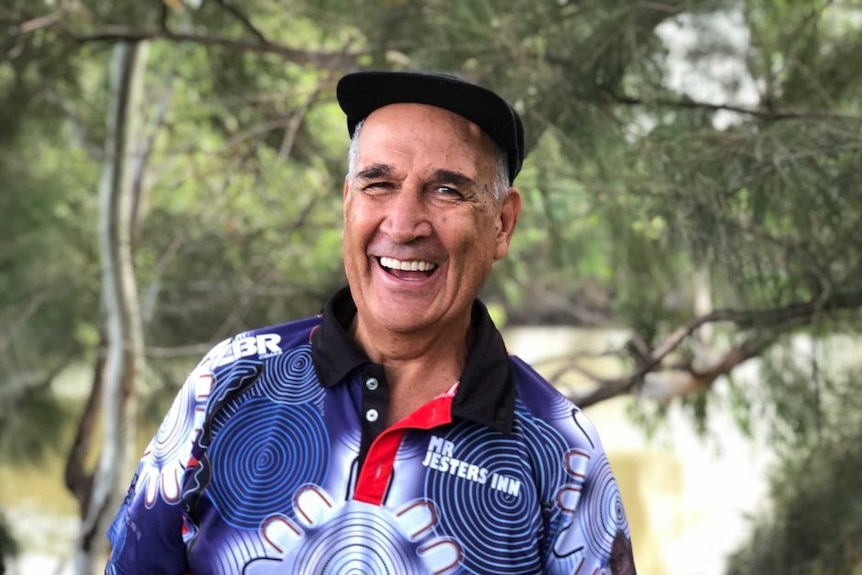 An Indigenous man in a cap smiles widely, looking at the camera.