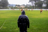Man wearing all black poncho stands with back to camera looking out toward rainy football field with players in distance