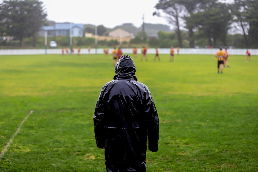Man wearing all black poncho stands with back to camera looking out toward rainy football field with players in distance
