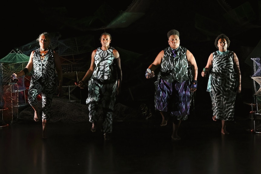 Four people in striking monochromatic printed outfits, walking side-by-side on stage against a blackened background.