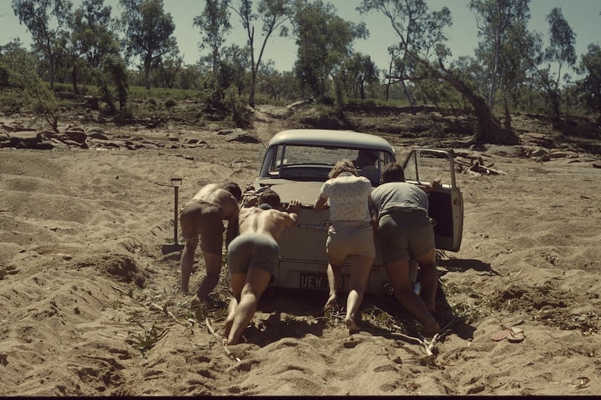 Four people pictured from behind pushing an old vehicle that is bogged. 